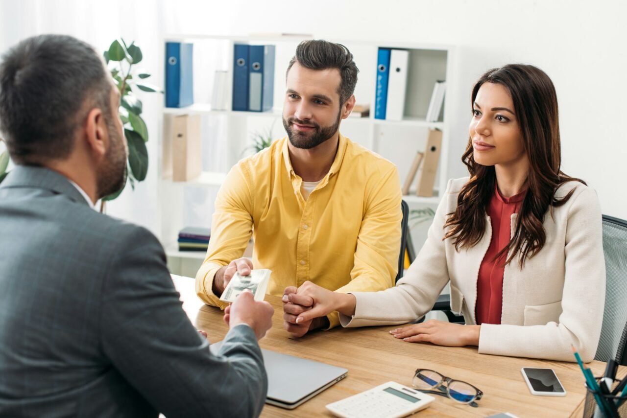 couple investors sitting at table and taking dollar banknotes from advisor in office